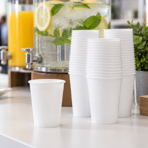 Stacks of white ribbed disposable plastic cups on a white counter, with beverage dispensers and greenery blurred in the background.