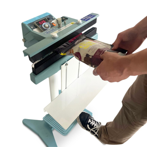 A light blue and white automatic food packaging sealing machine being operated by a person whose hands and legs are visible, sealing a packaged food item. The machine features a digital control display on top and a white flat sealing plate.