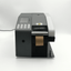A black and gray industrial label printer with a digital control panel, a roll of brown paper tape emerging from the output slot, and a power cord extending to the left, set against a plain white studio background.
