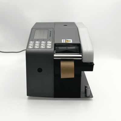 A black and gray industrial label printer with a digital control panel, a roll of brown paper tape emerging from the output slot, and a power cord extending to the left, set against a plain white studio background.