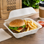 A gourmet burger with lettuce, tomato, and melted cheese on a sesame seed bun, served with golden french fries in a white foam take-out container on a wooden table. A small cup of ketchup, a paper napkin, a brown paper bag with 'GOOD FOOD GOOD MOOD' text, and a coffee cup are visible in the background.