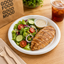 A plated salad with sliced grilled chicken breast, cucumber slices, tomato wedges, red onion, and a small cup of dressing on a white plate, set on a wooden table with a drink, utensils, and a paper bag in the background.