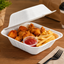 A white foam take-out container filled with fried chicken pieces and french fries, with a small cup of red ketchup, set on a wooden table with a fork, napkin, coffee cup, and plant in the background.