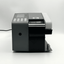 A compact industrial label printer or barcode scanner with a dark gray and black body, featuring a control panel with buttons on the left, a paper slot in the center, and a light gray cover on the right, photographed on a light gray studio background.