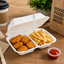 A white foam take-out container holding golden-brown fried chicken nuggets and french fries on a wooden table, with a small cup of red ketchup, a takeaway coffee cup, napkins, and a brown paper bag printed with 'GOOD FOOD GOOD MOOD'.