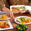 Person pointing at a plate of food with various dishes including salad, fries, and waffles on a wooden table.