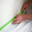 A person's hands are using green masking tape on a white surface, likely a wall, with a white garment laid out on the surface for protection.