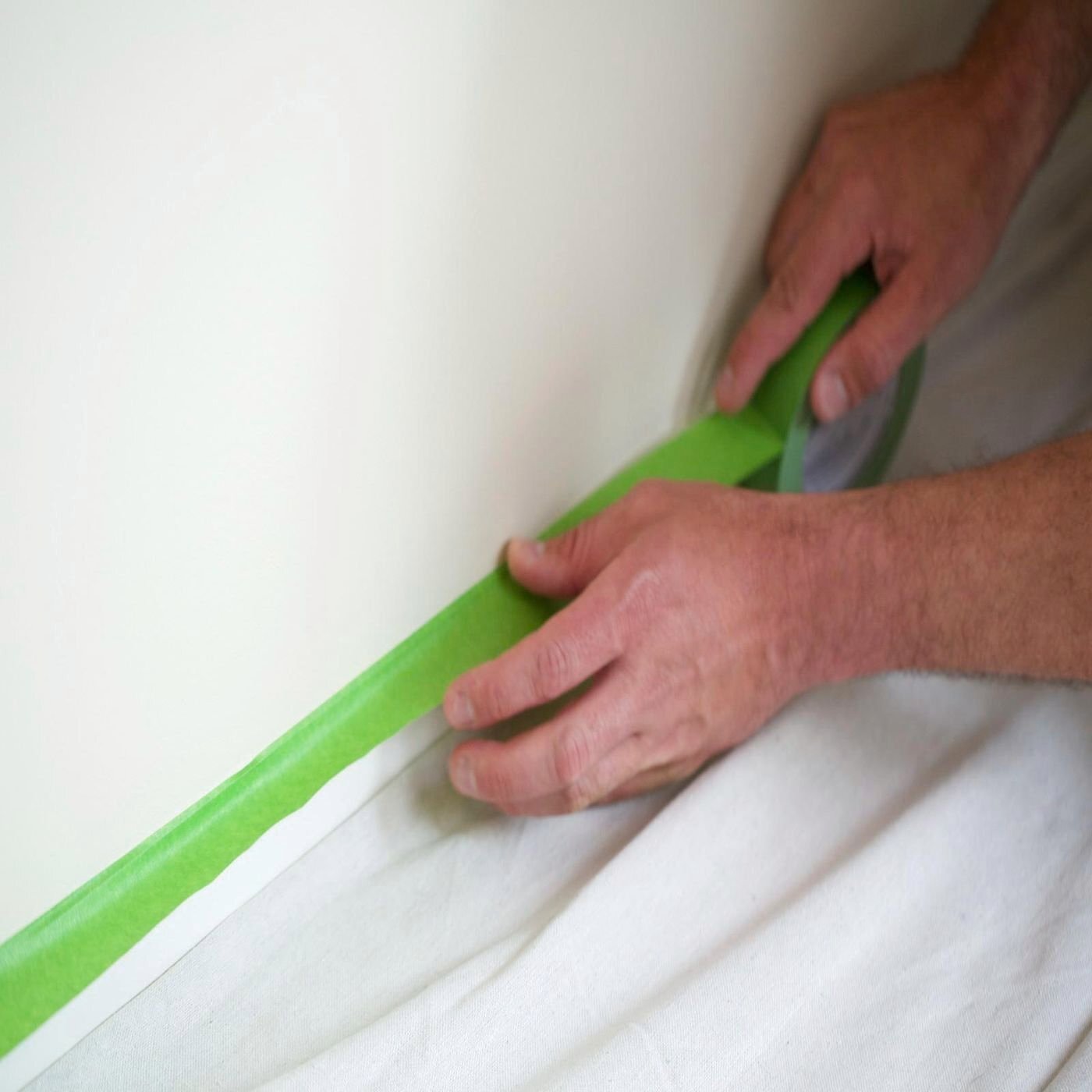 A person's hands are using green masking tape on a white surface, likely a wall, with a white garment laid out on the surface for protection.