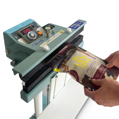 A teal Xenc brand manual heat sealer machine being used by a person to seal a bag of gourmet chocolate chips. The machine features a digital temperature display, adjustment knobs, and a black mesh conveyor belt, with the person's hands holding the bag under the sealer against a clean white background.