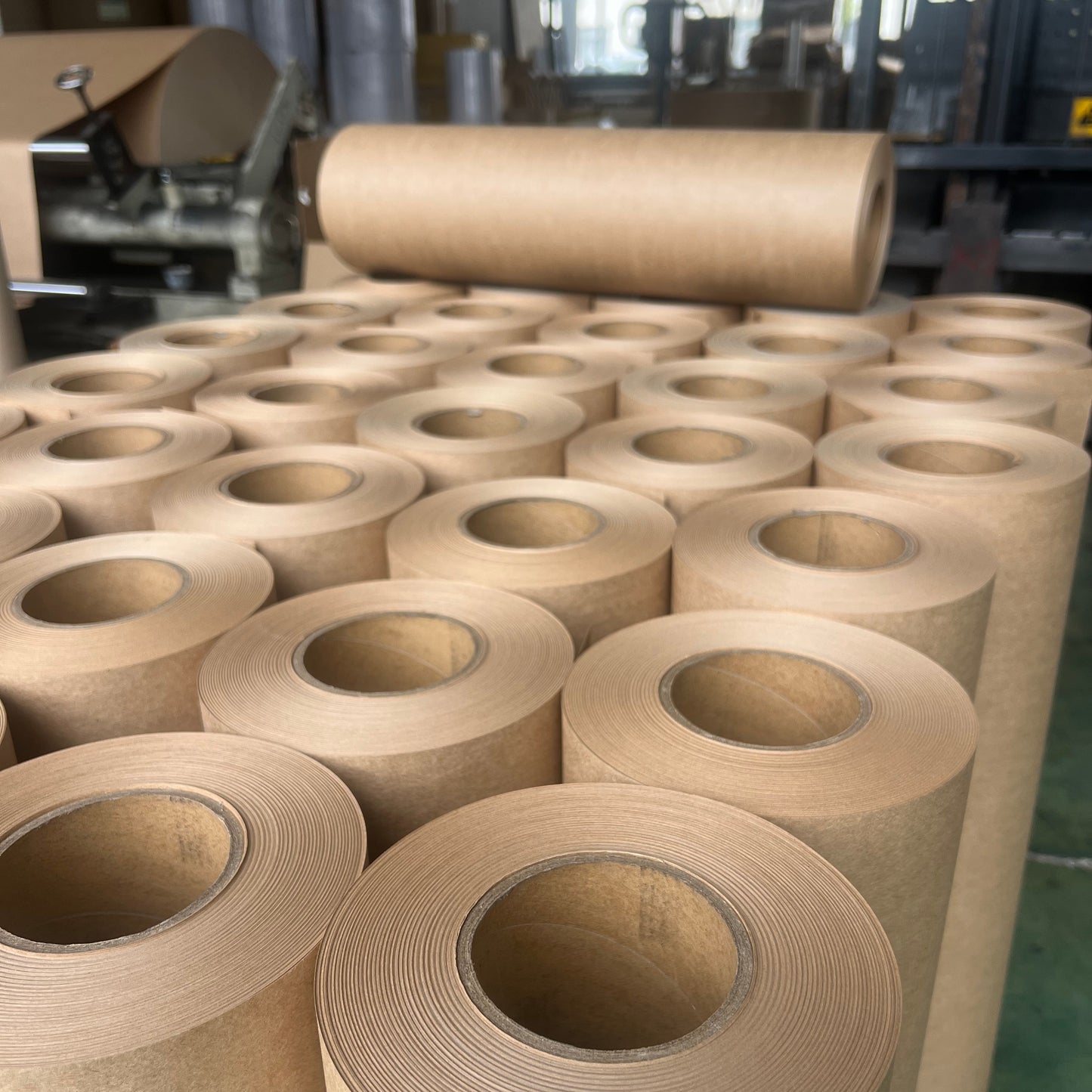 Multiple rolls of tan kraft paper or cardstock wound around cylindrical cores, arranged in rows on a factory floor with industrial machinery visible in the background.