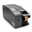 A black and white industrial barcode scanner with a digital display screen and numeric keypad, shown with a roll of brown paper tape emerging from the port, on a white background.