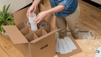 Person packing glass items into a cardboard box on a wooden table.
